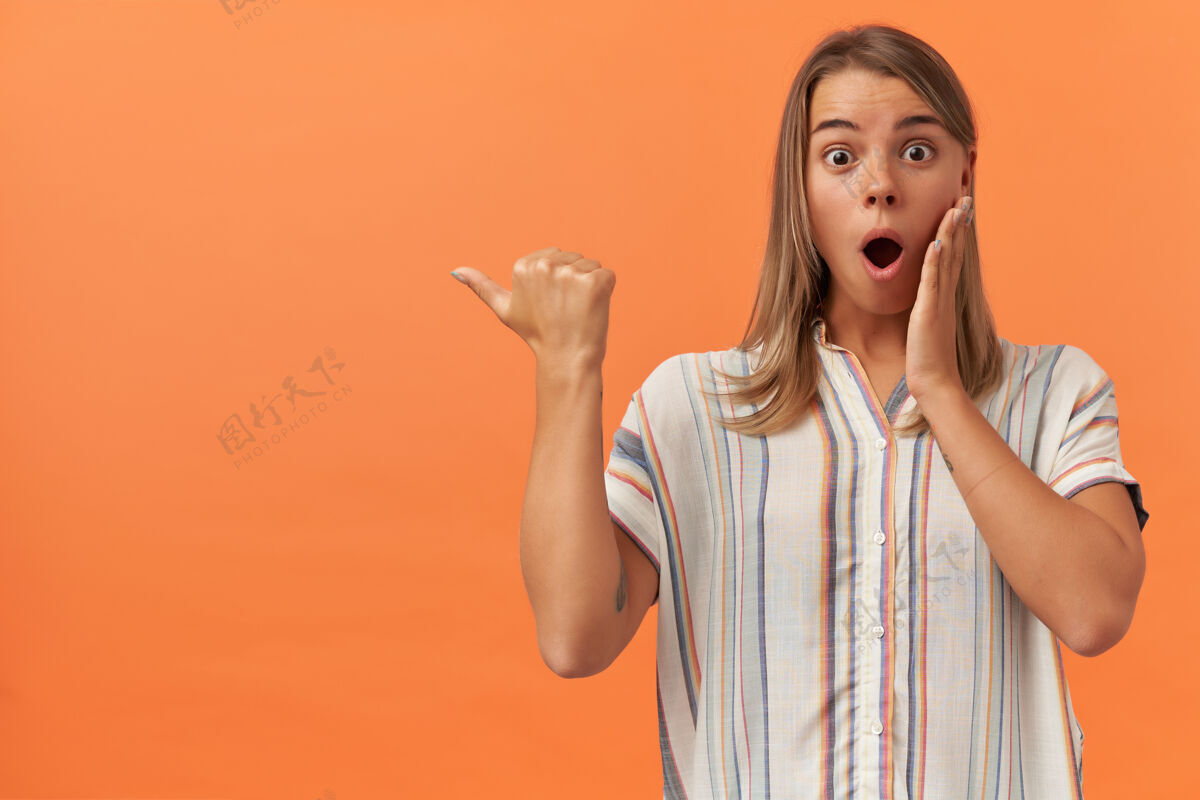 女人一位穿着条纹衬衫的年轻女士惊讶地把手放在脸颊上 用拇指指着被隔离在橙色墙壁上的版面边惊人指向女性