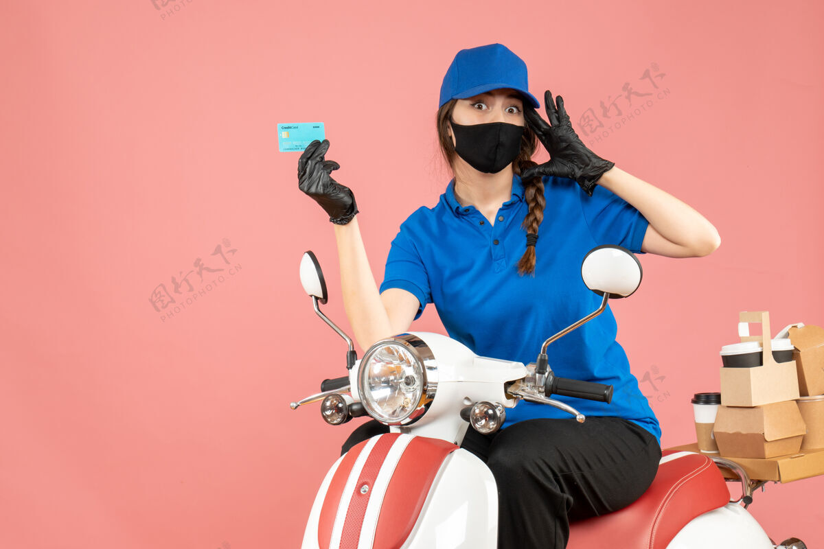 桃顶视图混乱的女快递员戴着医用面罩和手套坐在滑板车上拿着银行卡在粉彩桃色背景下传递订单车辆举行交付