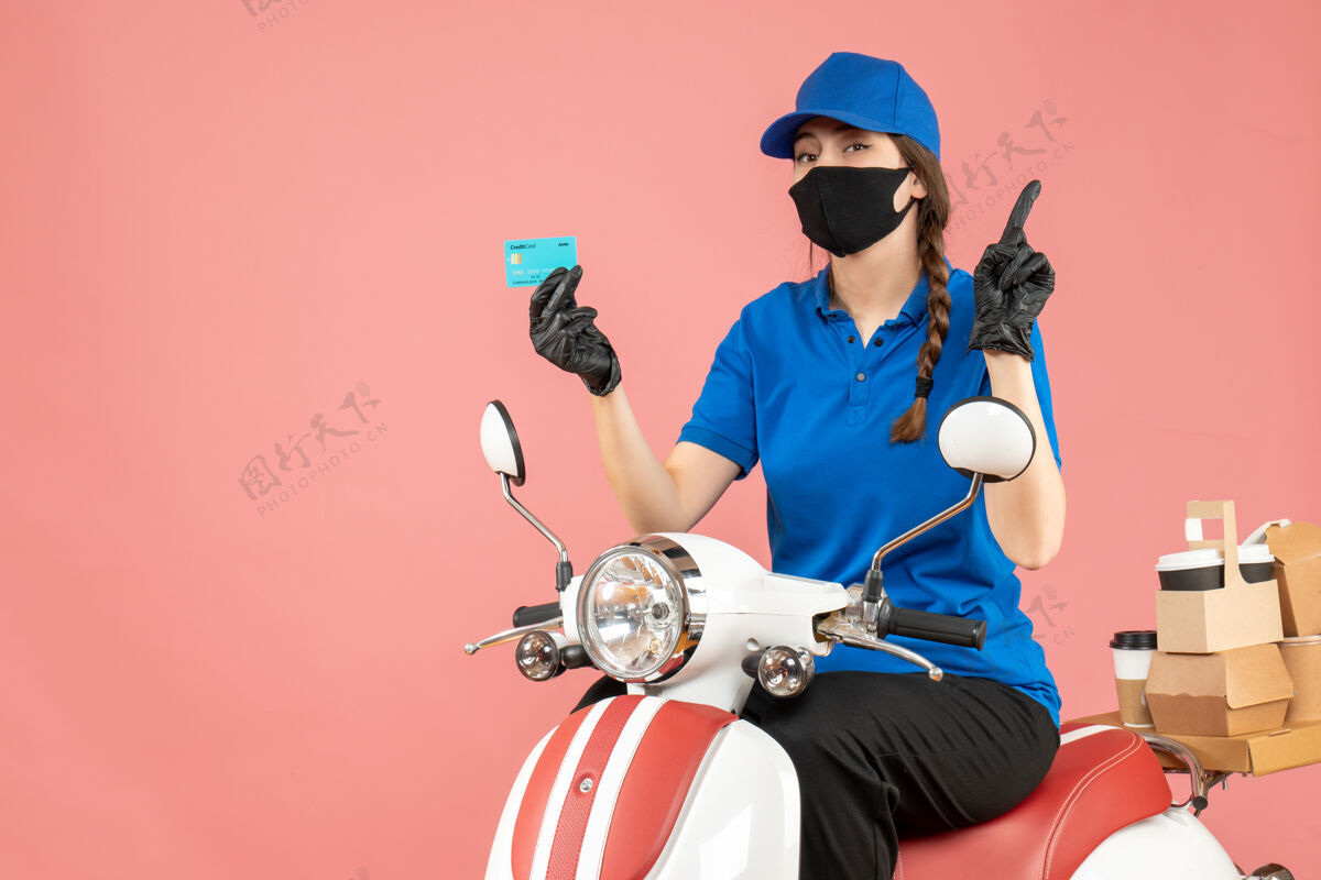 背景俯视图：戴着医用面罩和手套的女快递员坐在滑板车上 手里拿着银行卡 在粉彩桃色背景上向上传递订单交付持有手套