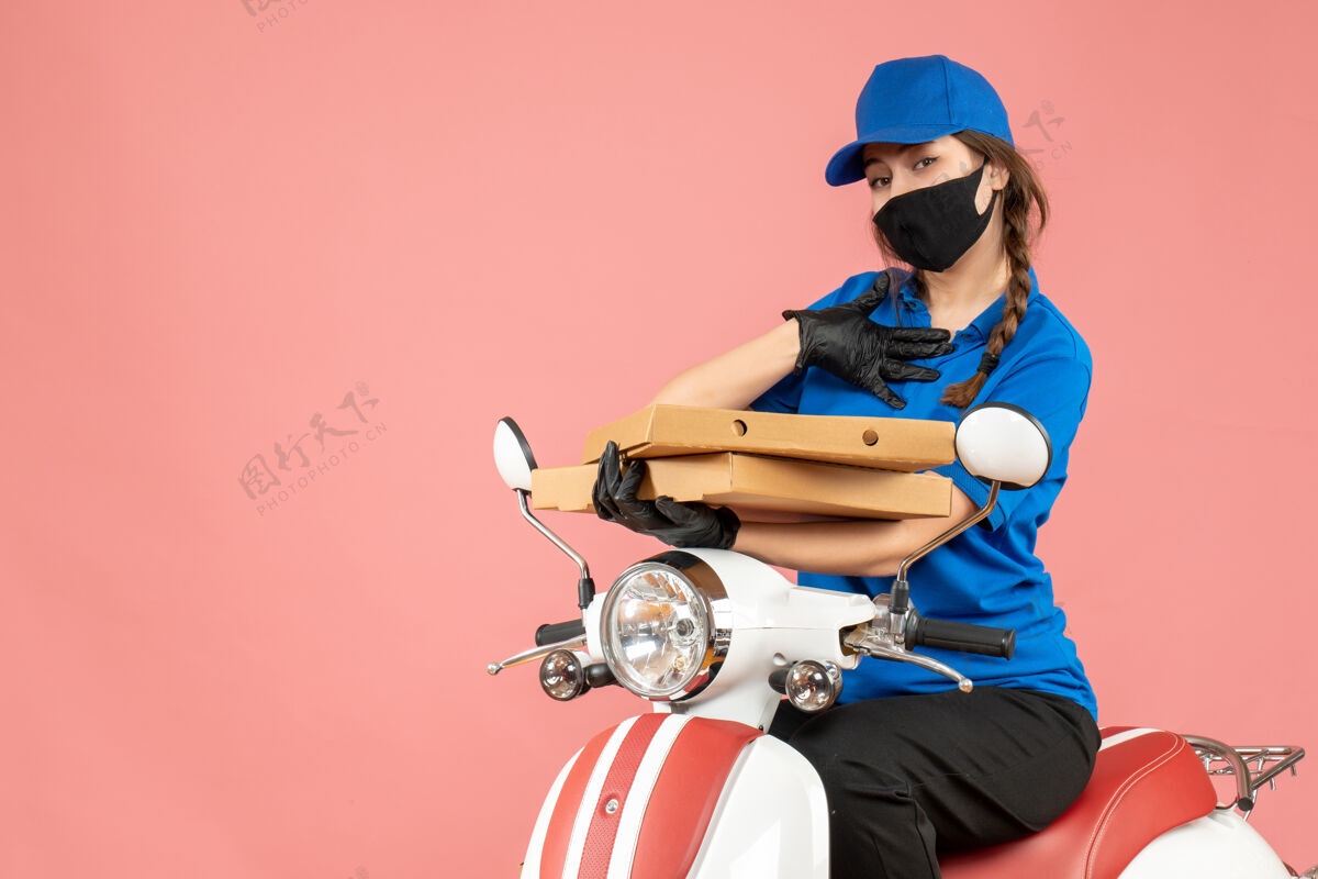 手套前视图年轻的女快递员戴着医用面罩和手套坐在滑板车上传递粉彩桃背景的命令背景粉彩桃