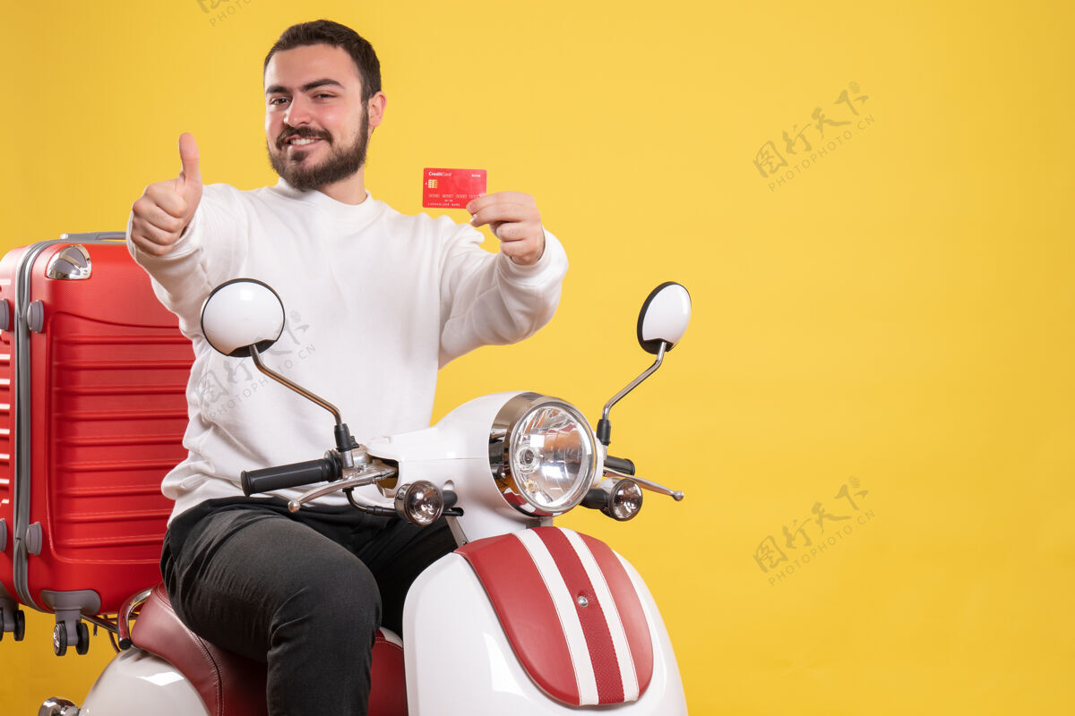 举行正面图是一个年轻的面带微笑的旅行男子坐在摩托车上 手提箱上拿着银行卡 在孤立的黄色背景上做着“ok”的手势车辆好手势