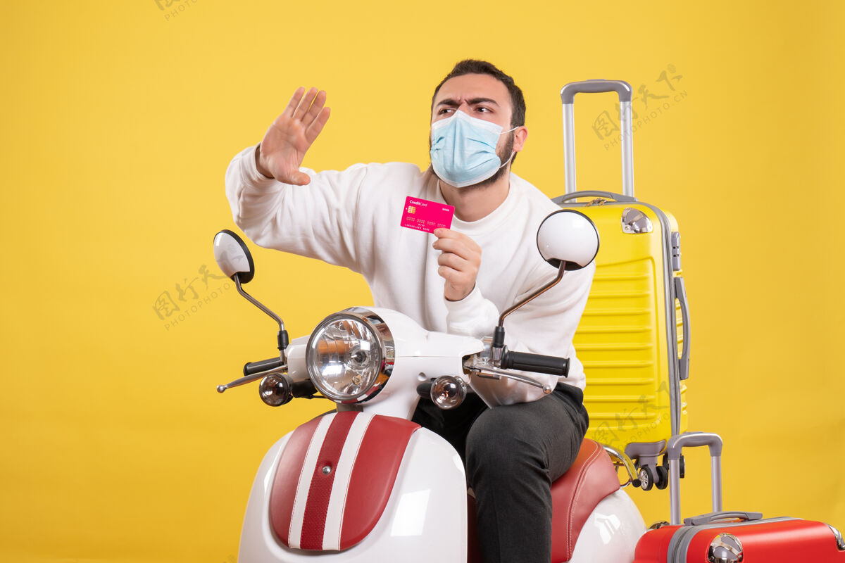 人旅行概念的正面图 戴着医用面罩的年轻紧张的家伙坐在摩托车上 黄色手提箱放在上面 手里拿着银行卡医疗手提箱坐着