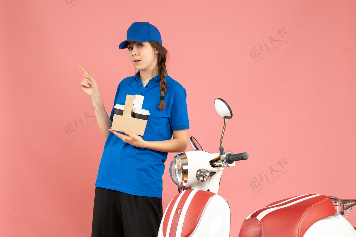 专业前视图好奇的信使女孩站在摩托车旁边拿着咖啡指着粉红色的背景咖啡站粉彩