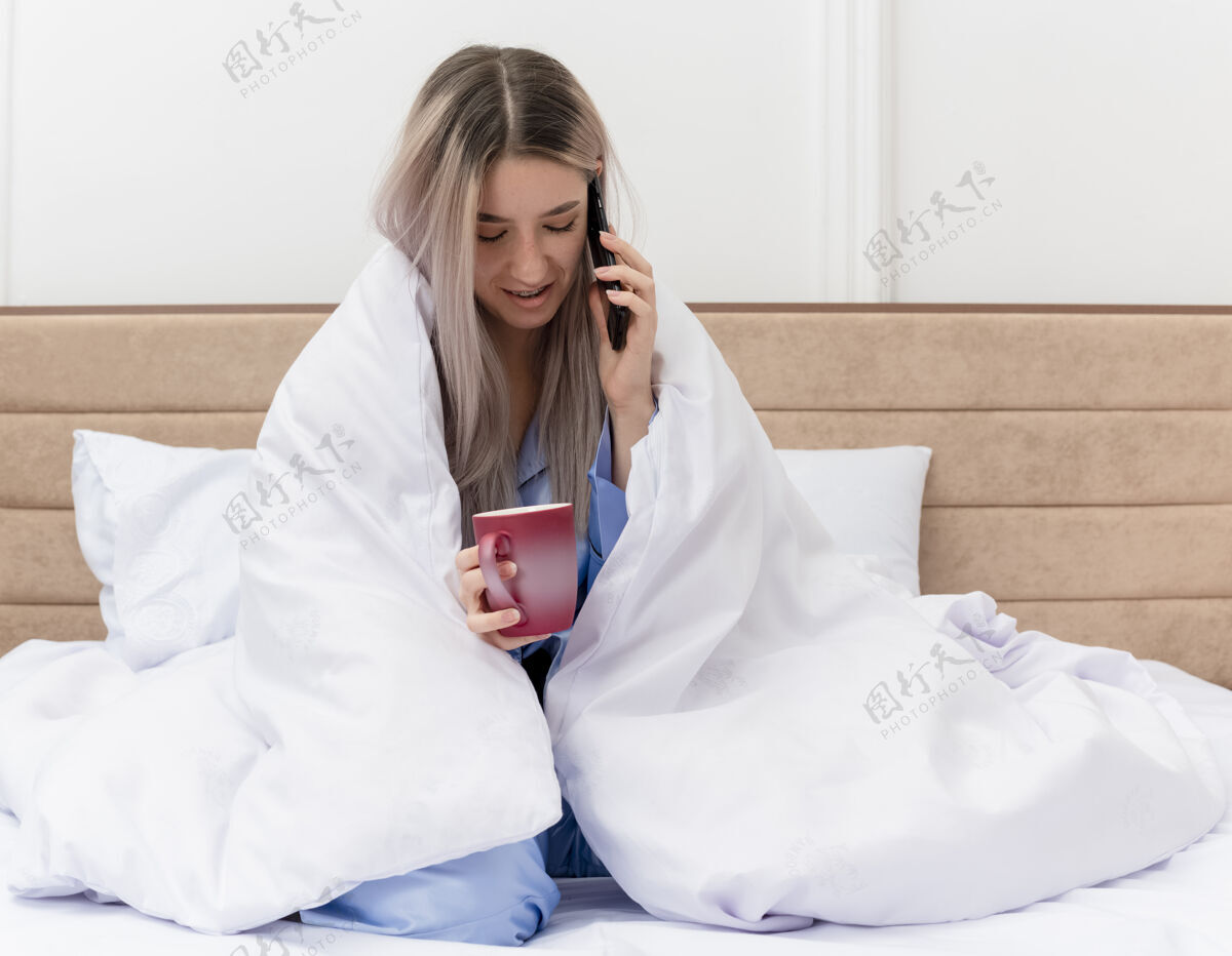 电话穿着蓝色睡衣的年轻美女坐在床上 裹着毯子 端着咖啡 在卧室里用手机聊天杯子室内背景