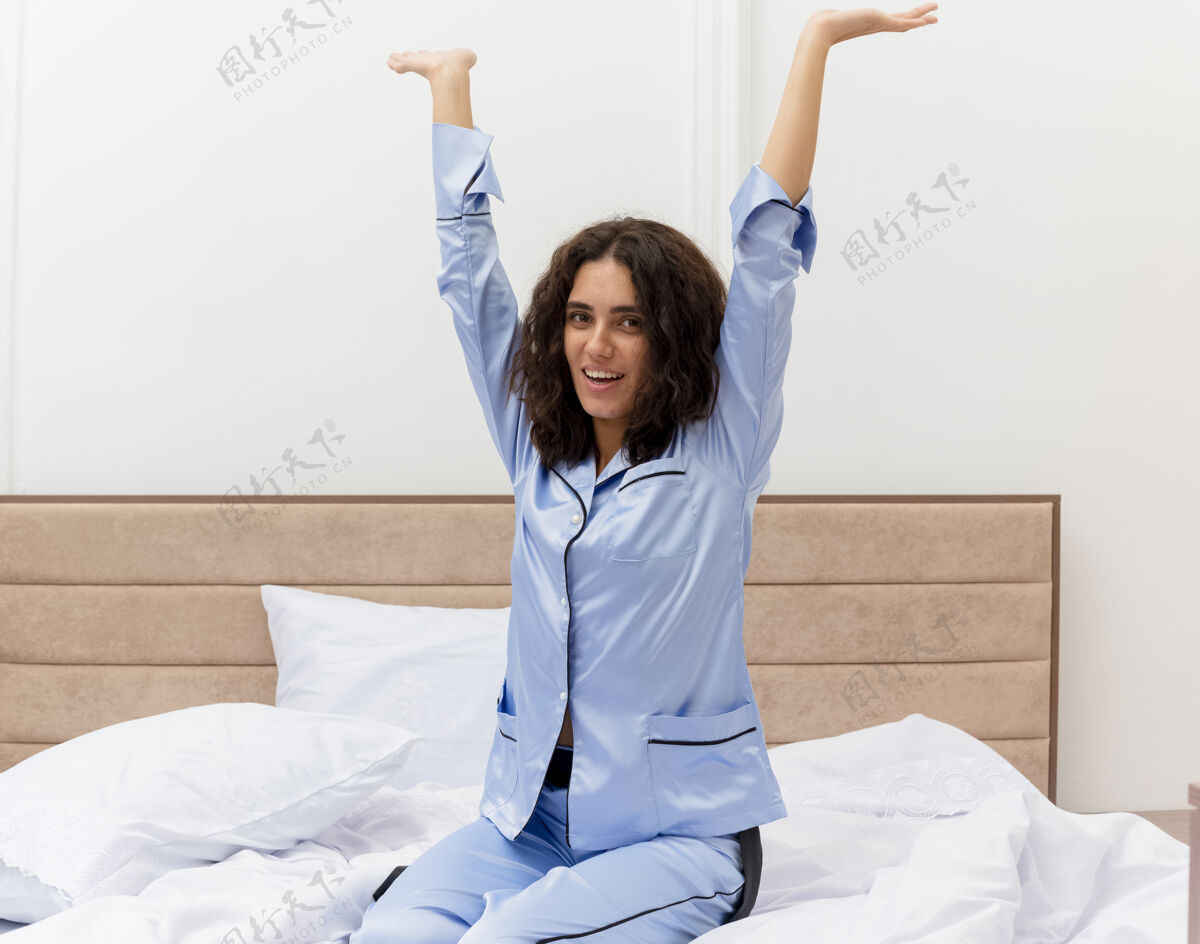 年轻穿着蓝色睡衣的有趣年轻漂亮女人坐在床上快乐而积极地享受周末在卧室里积极快乐蓝色