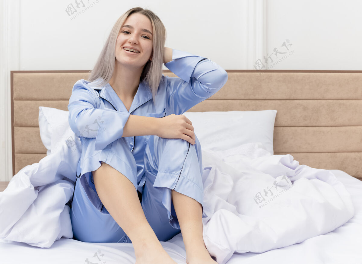 漂亮穿着蓝色睡衣的年轻漂亮女人坐在床上快乐而积极地微笑着 在卧室里休息灯光睡衣蓝色