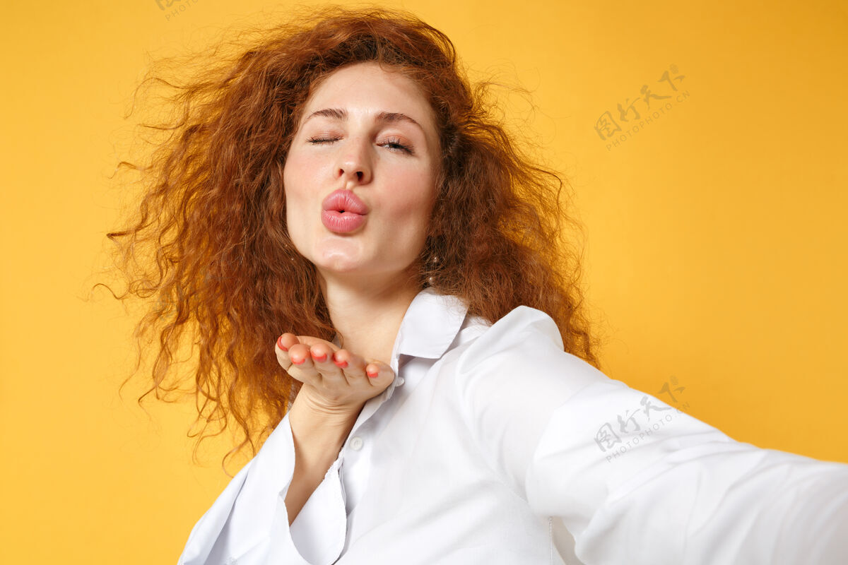 人物特写镜头：穿着白衬衫的年轻红发女子在黄橙色的墙上孤零零地摆姿势女性秀亲吻