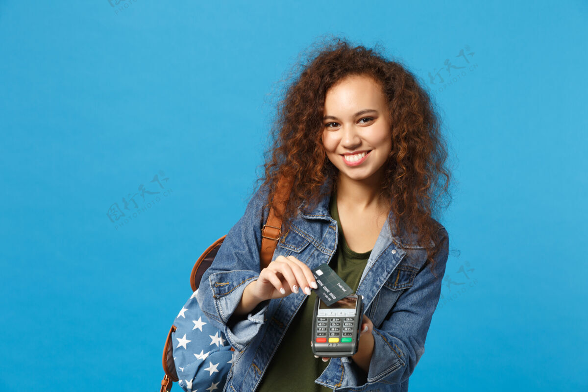 信贷年轻的非洲裔美国女孩十几岁的学生在牛仔服装 背包举行信用卡隔离在蓝色的墙上货币非接触学生
