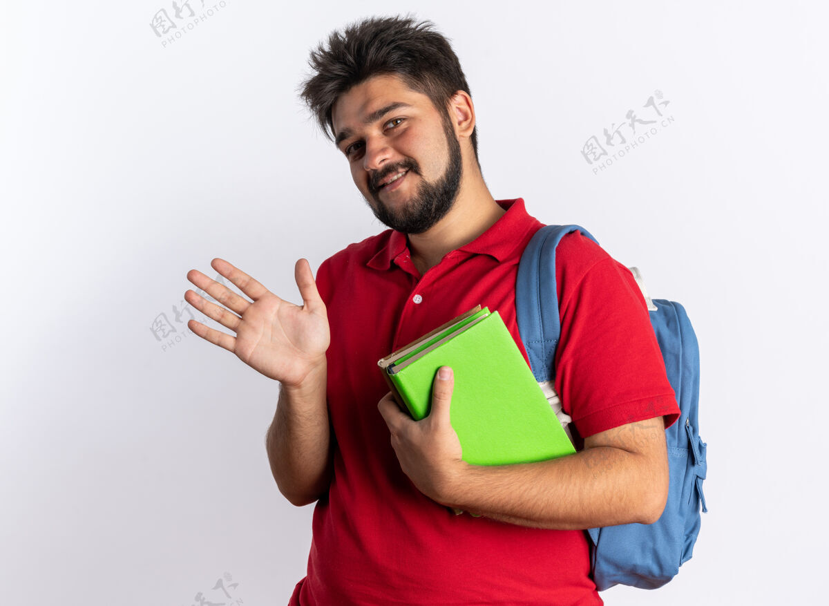 挥手年轻的留着胡子的学生 穿着红色马球衫 背着书包 拿着笔记本 快乐而愉快地微笑着 手站在白色的墙上挥舞着年轻人背包马球