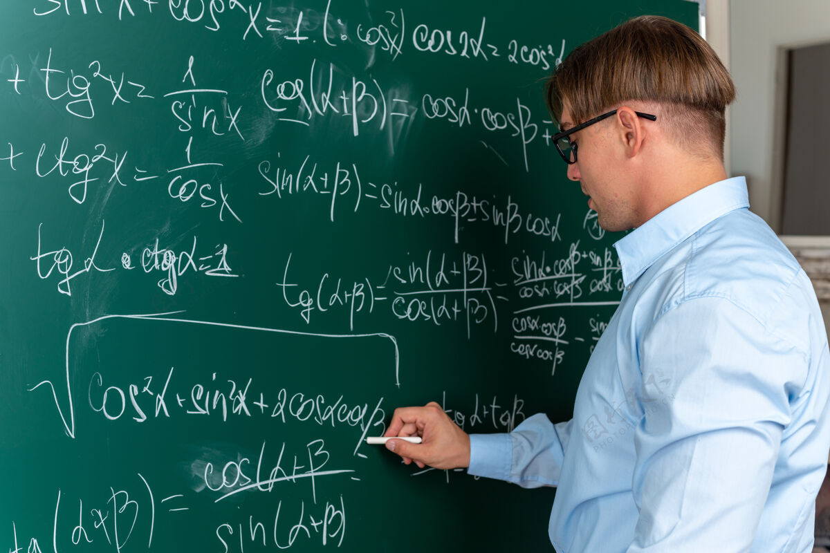 教室戴着眼镜的年轻男老师站在黑板旁 在黑板上写数学公式讲解课 在教室里显得自信满满写作穿解释