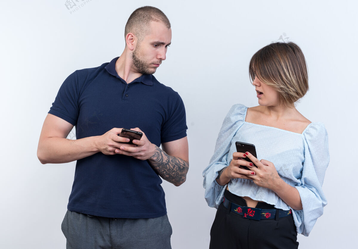衣服年轻漂亮的一对穿着休闲服的男女拿着智能手机站在白色的墙壁上困惑地看着对方女人站立人