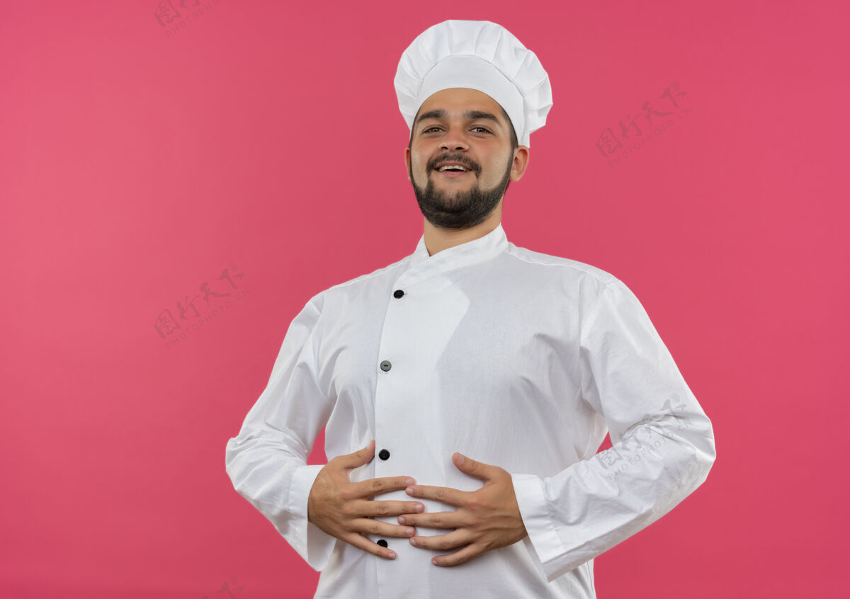 男性身着厨师制服的快乐年轻男厨师将手放在腹部隔离在粉色墙壁上 并留有复制空间快乐制服腹部