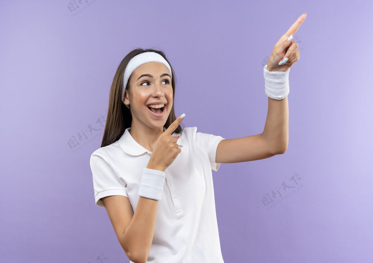 漂亮快乐漂亮的运动女孩戴着头带和腕带指着隔离在紫色墙上的一边运动欢乐女孩