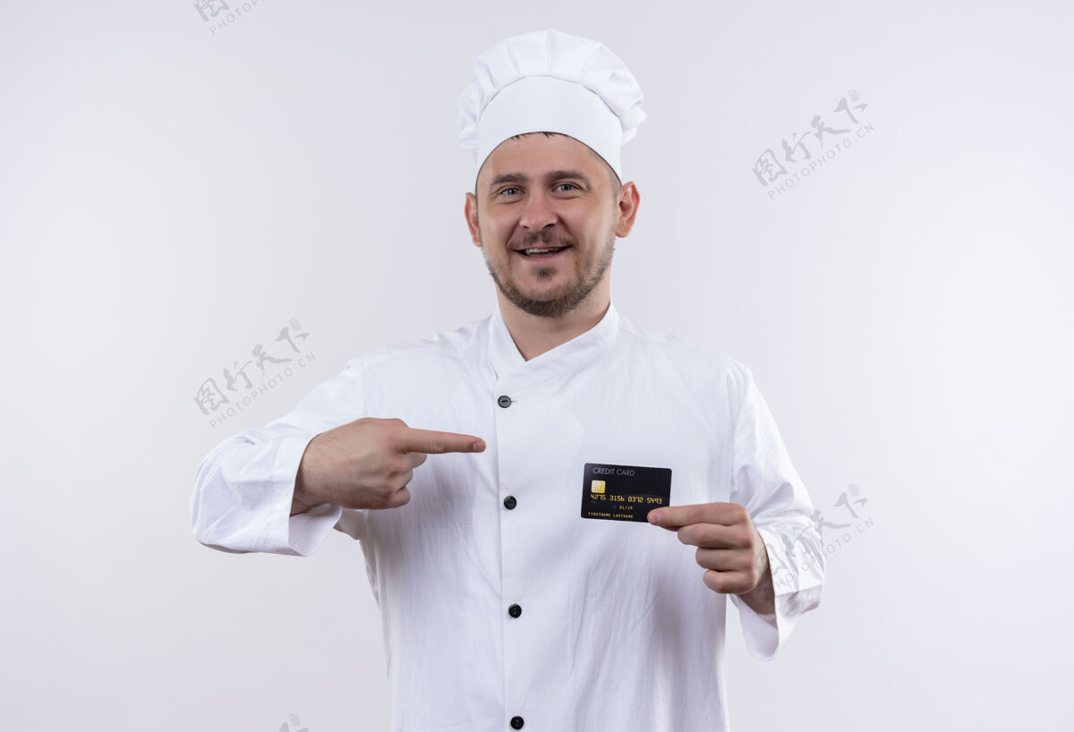 指向身着厨师制服的年轻帅气的厨师手拿着信用卡 指着隔离在白墙上的那张英俊制服快乐