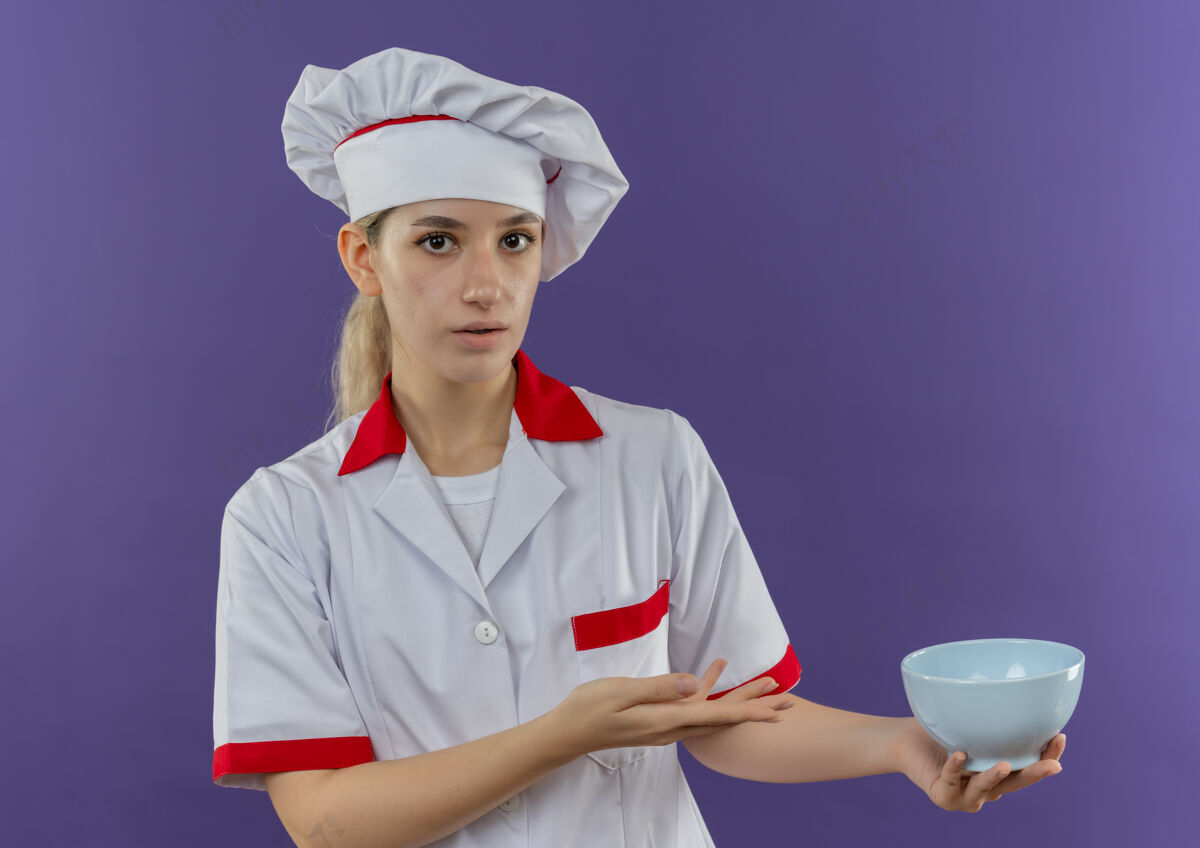 制服令人印象深刻的年轻漂亮厨师在厨师制服举行 并用手指着碗隔离在紫色的墙壁拿着年轻手