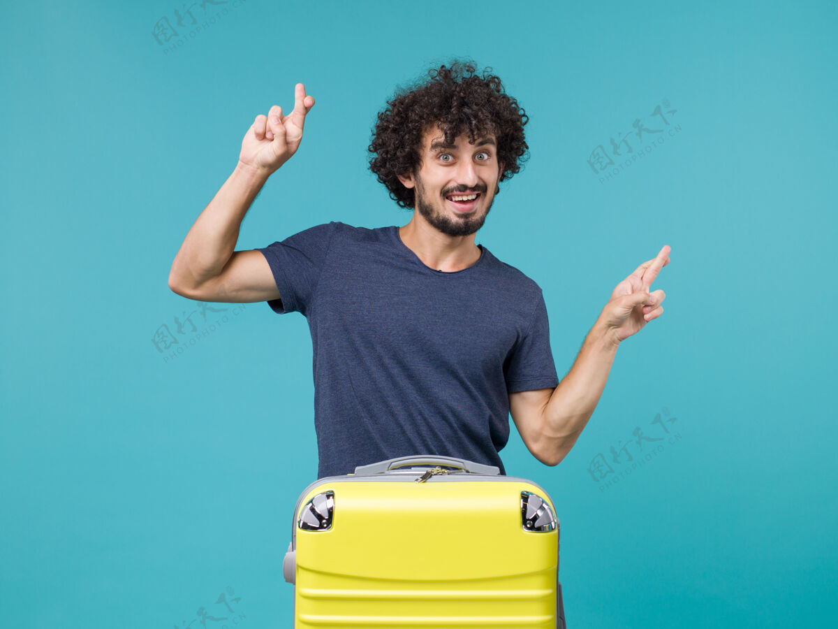旅行带着黄色手提箱的度假男人把手指交叉在蓝色手提箱上十字架黄色手指