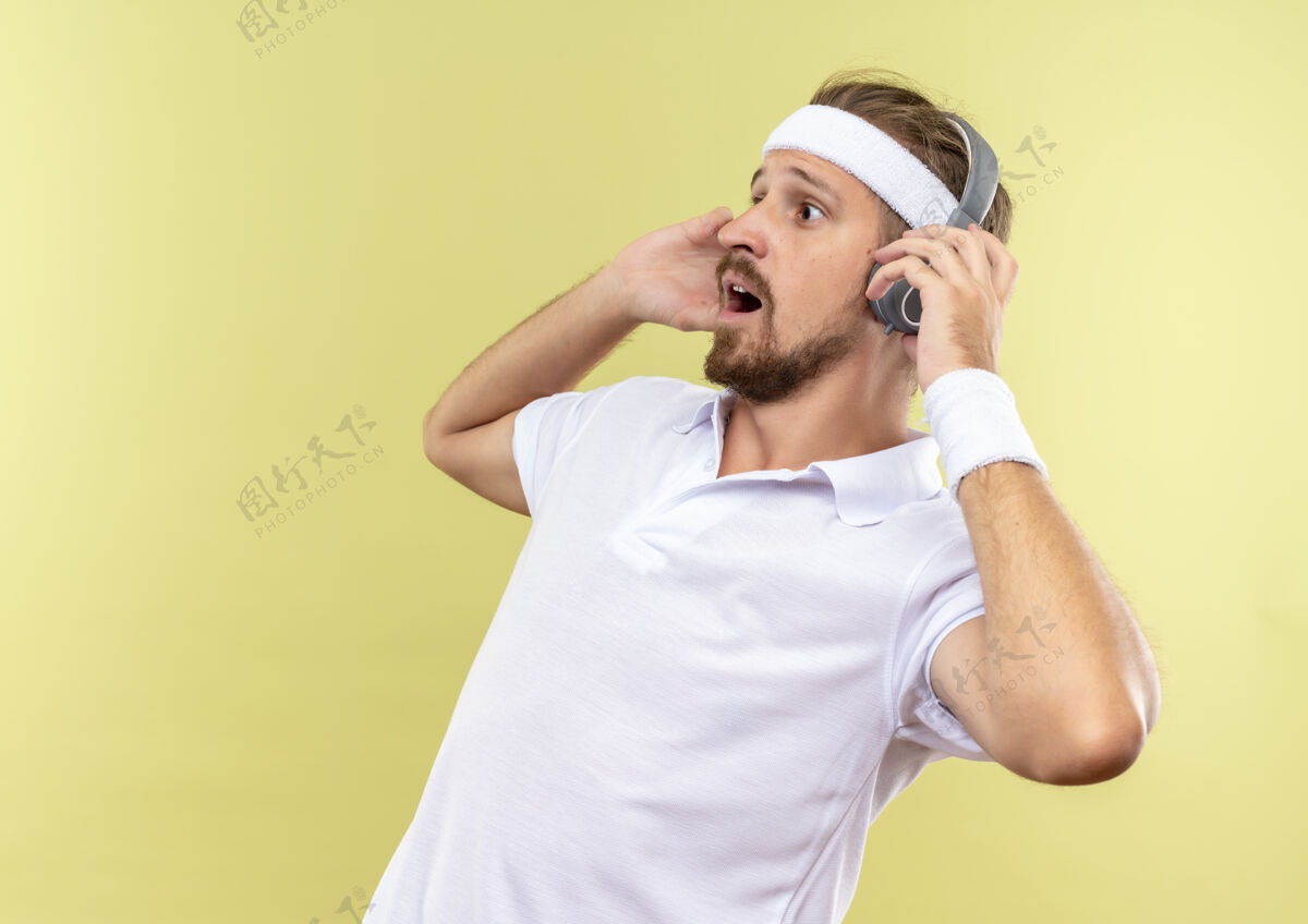 头带印象深刻的年轻英俊的运动男子戴着头带和腕带和耳机看着一边用手戴着耳机隔离在绿色墙壁上的复制空间手年轻壁板