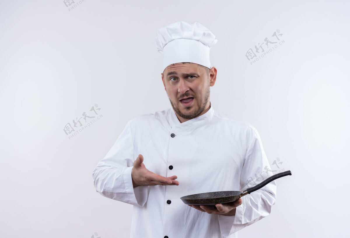 制服身着厨师制服的年轻帅哥自信地拿着 指着隔离在白墙上的煎锅平底锅拿着指着