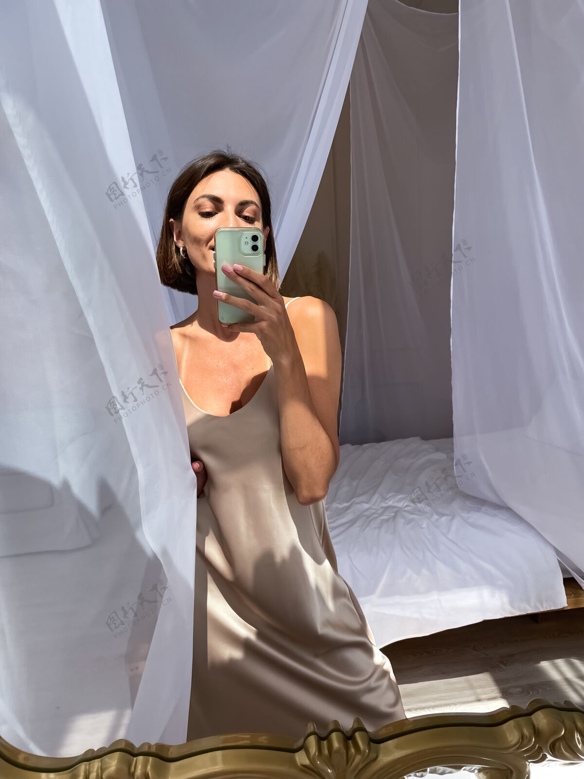 假期穿着浪漫米色丝绸连衣裙的健康晒黑女人在家里对着镜子在电话里自拍电话温暖自画像