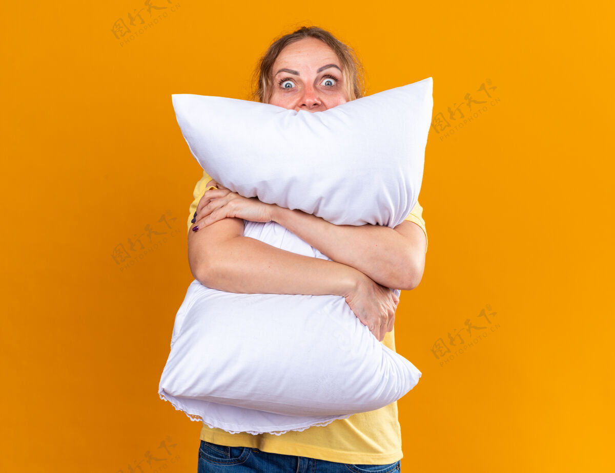 痛苦一个不健康的女人穿着黄色衬衫 患流感和感冒 感觉不舒服 抱着枕头 害怕站在橙色的墙上女人流感害怕