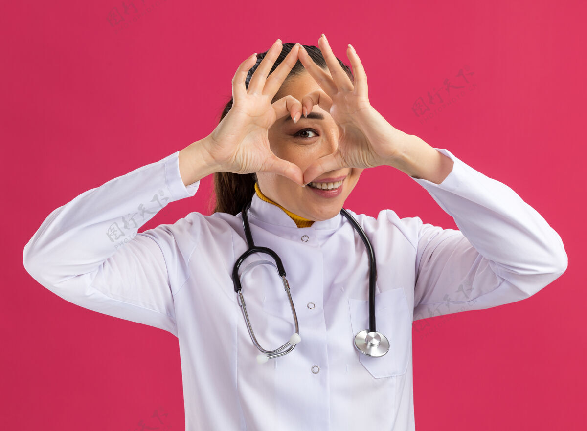 手势身穿白色药衣的年轻女医生脖子上戴着听诊器做着心脏手势 手指透过手指微笑着站在粉色的墙上心脏外套女人