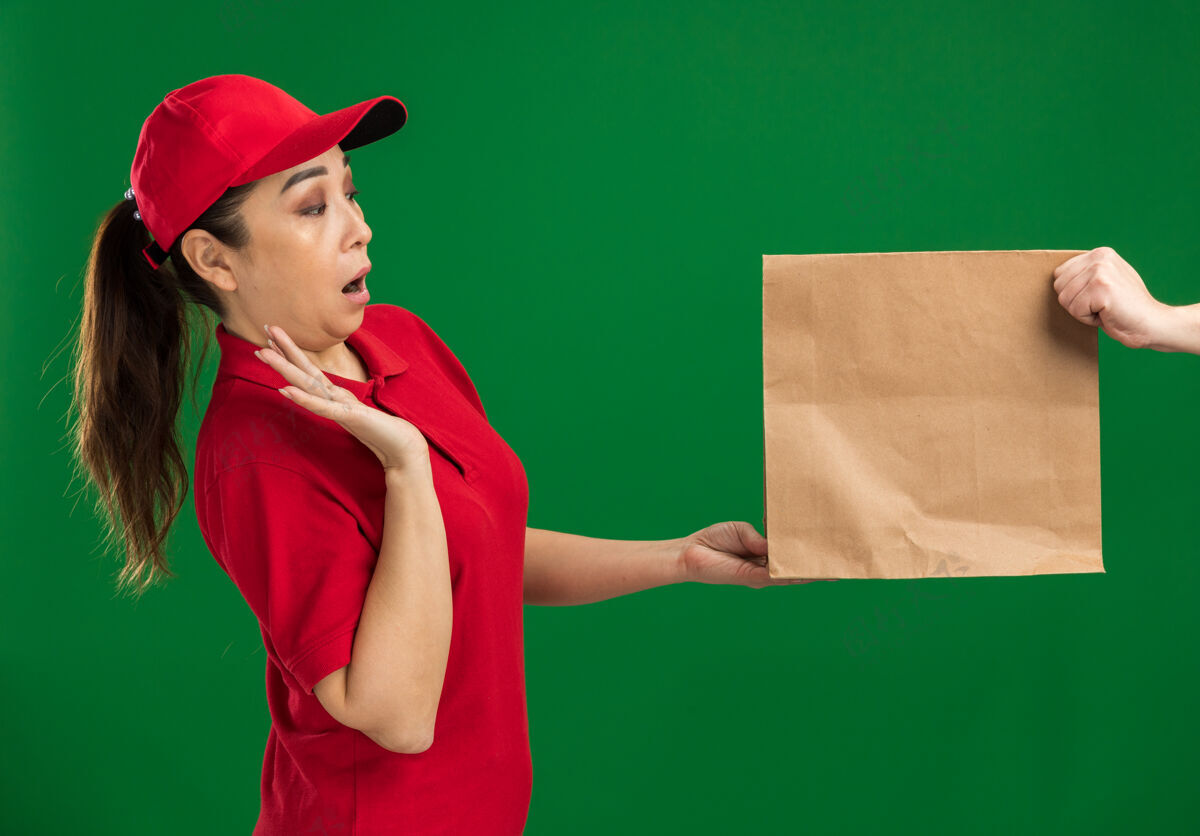 女孩年轻的送货女孩穿着红色制服 戴着帽子 拒绝接受纸包裹 看着它惊讶地站在绿色的墙上惊喜帽子接收