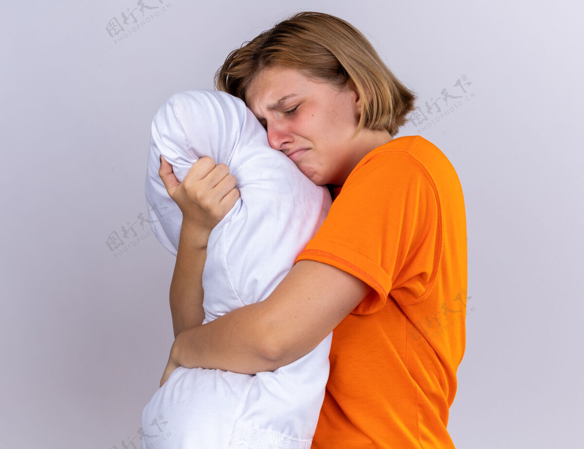 枕头心烦意乱的不健康的年轻女子穿着橙色t恤 抱着枕头 感觉不舒服 患流感 站在白色的墙上痛哭女人痛苦心烦