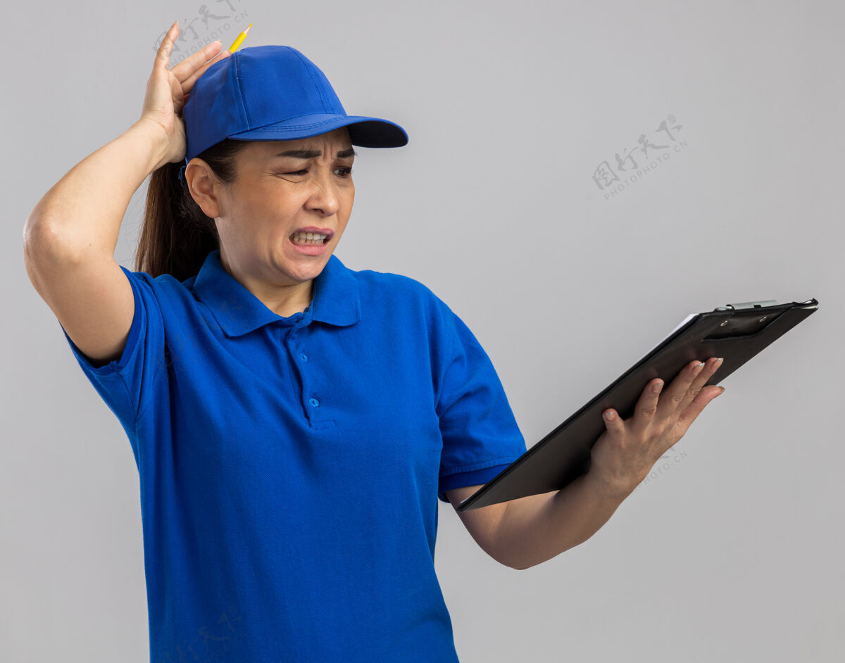交货穿着蓝色制服 戴着帽子的年轻女送货员拿着剪贴板 困惑地看着它 不高兴地把手放在头上 因为她站在白墙上犯了错误制服错误立场