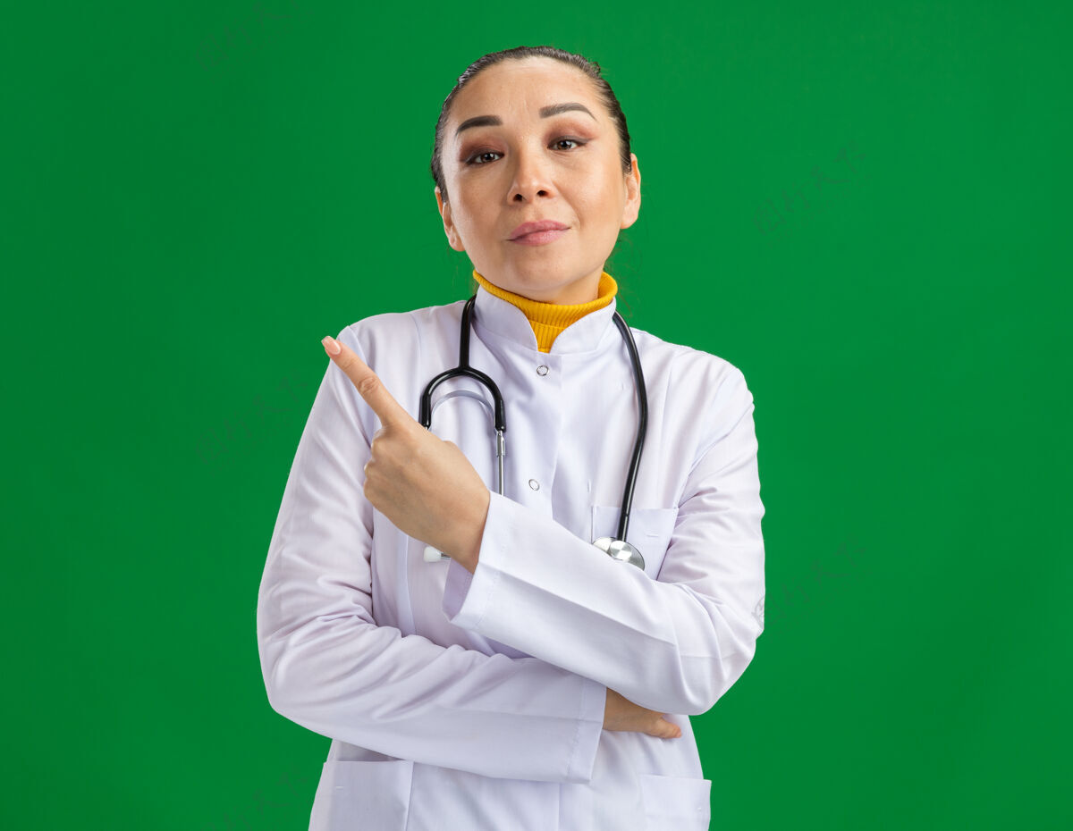 医生年轻的女医生穿着白药衣 脖子上戴着听诊器 看起来很自信 用食指指着绿墙那边年轻人站指向