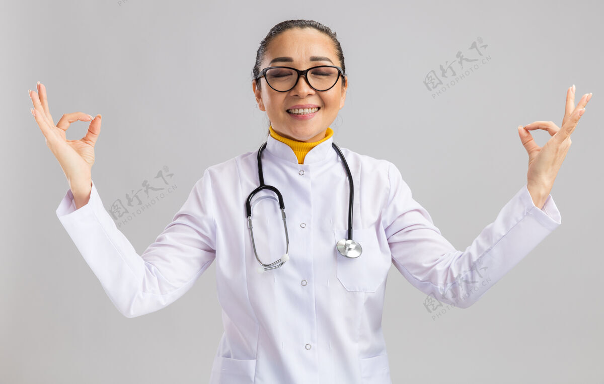 脖子年轻的女医生穿着白色的医用外套 脖子上戴着听诊器 手指站在白色的墙上 做着冥想的姿势女人站外套