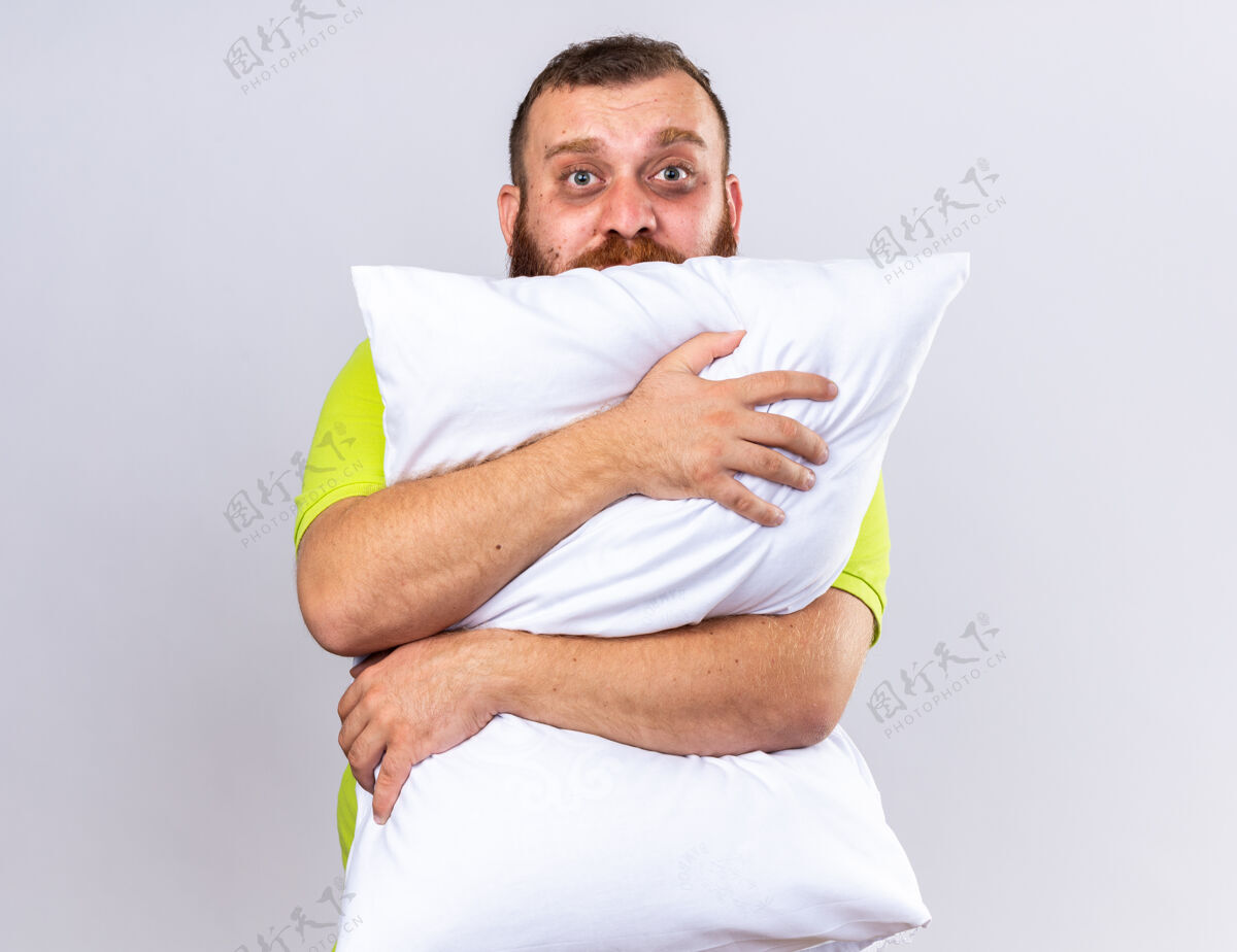 枕头不健康的胡子男人穿着黄色马球衫感觉不舒服拥抱枕头与悲伤的表情站在白墙不健康生病马球