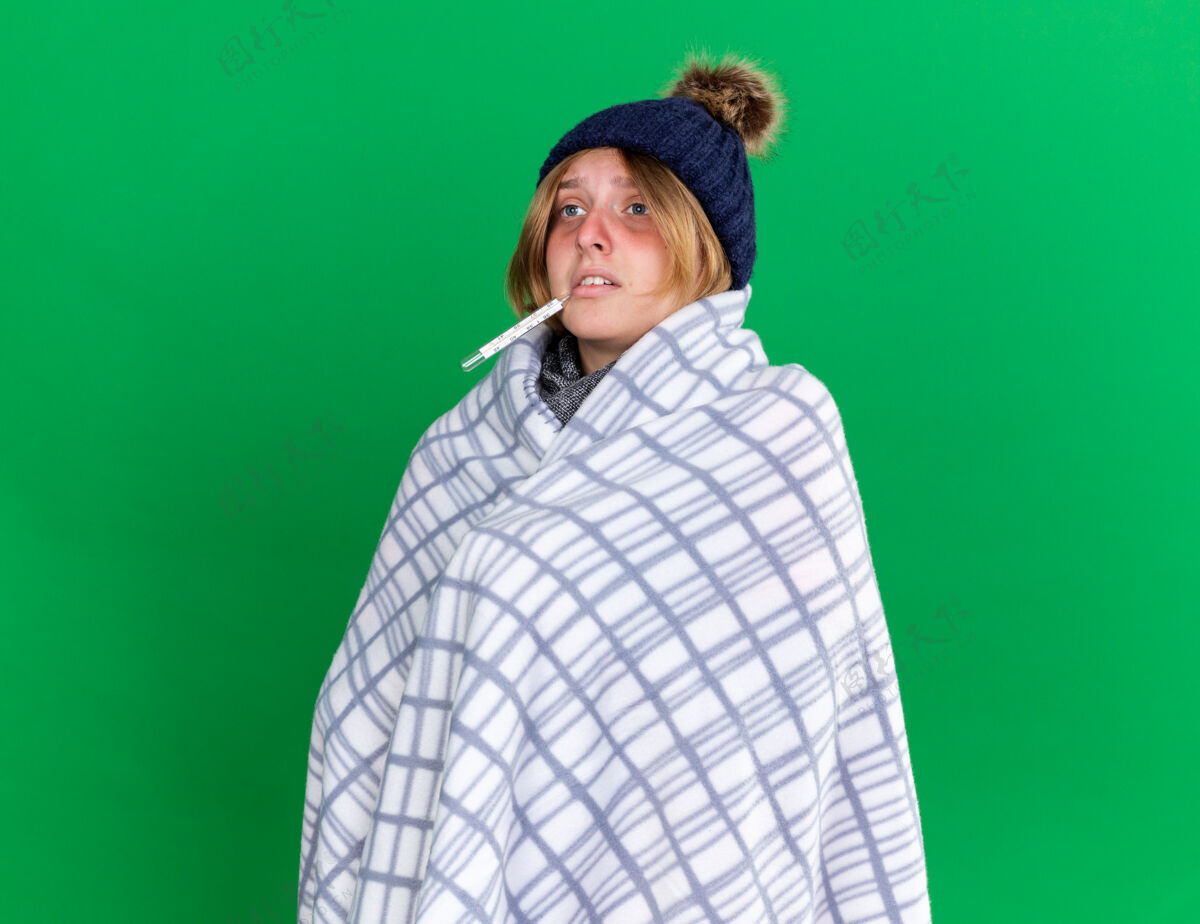 穿不健康的年轻女子裹着毯子戴着帽子用体温计测量体温患流感发烧站在绿色的墙上发烧痛苦体温计