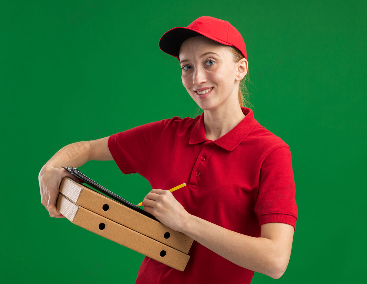 微笑身穿红色制服 头戴鸭舌帽 拿着披萨盒 拿着空白纸和铅笔的剪贴板的年轻送货女孩自信地站在绿色的墙上送货制服盒子