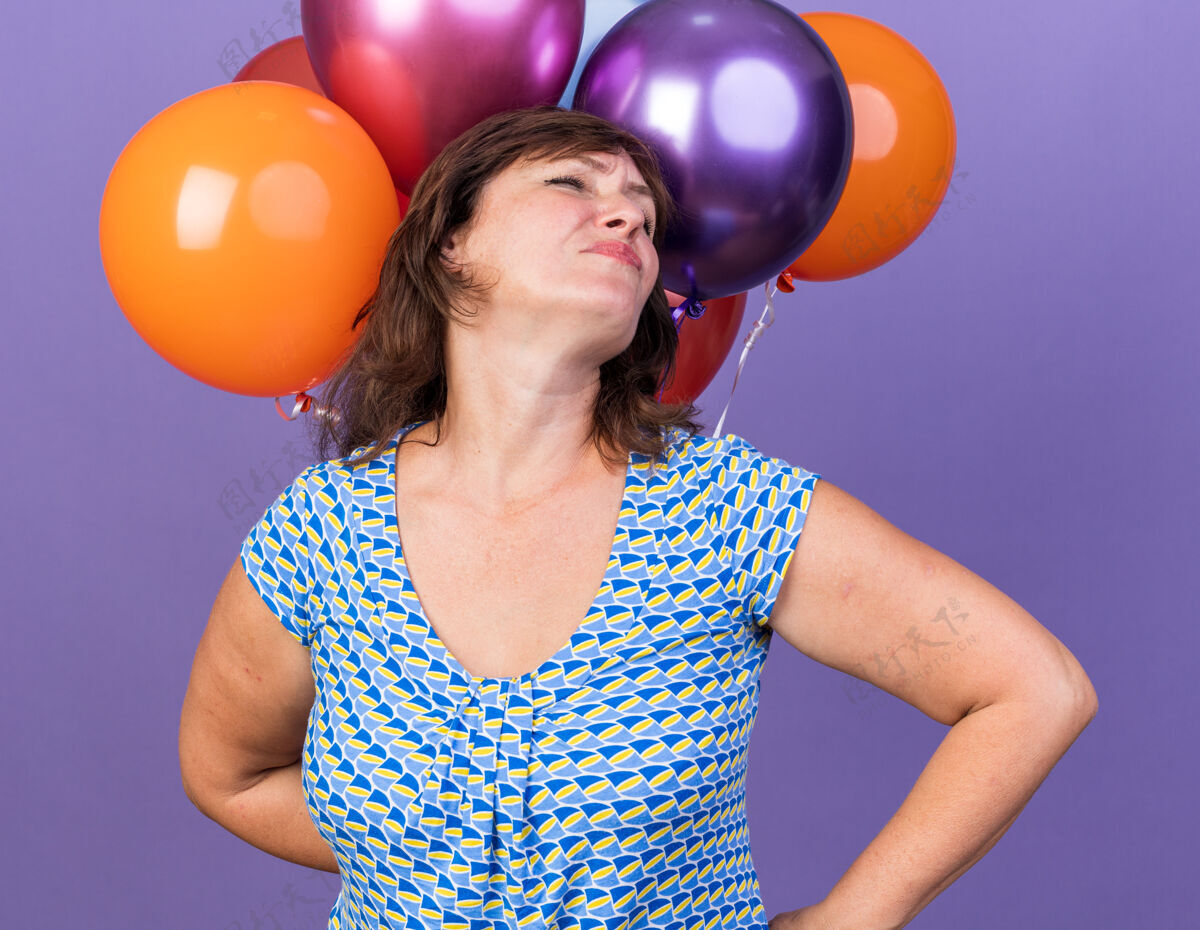 中间快乐的中年妇女带着一束五颜六色的气球微笑自信地站在紫色的墙上庆祝生日派对立场庆祝束