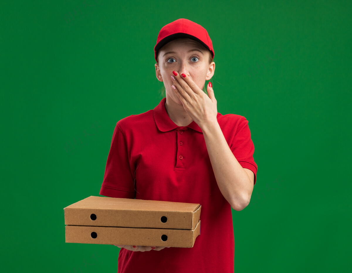 披萨穿着红色制服 戴着帽子的年轻送货女孩拿着披萨盒 手捂着嘴站在绿色的墙上 震惊不已女孩送货制服