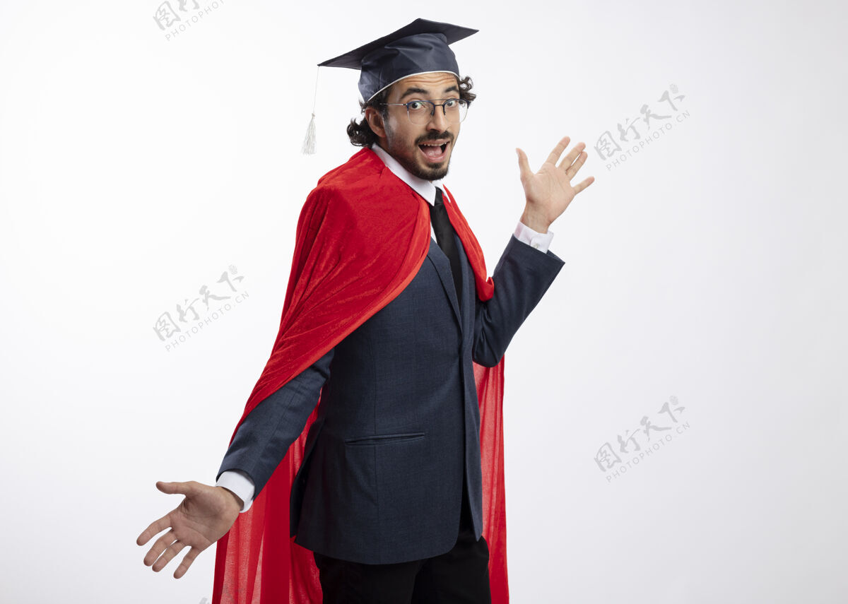 穿快乐的年轻白种人超级英雄 戴着眼镜 身穿红色斗篷 戴着毕业帽的西装 双手张开 站在一旁光学年轻持有