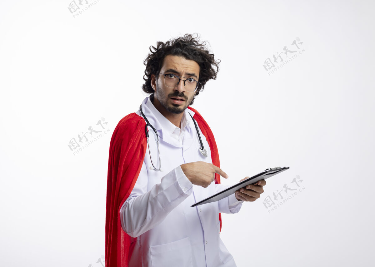 斗篷无知的年轻白种人超级英雄戴着眼镜 穿着医生制服 披着红色斗篷 脖子上戴着听诊器 拿着并指着剪贴板穿红色周围