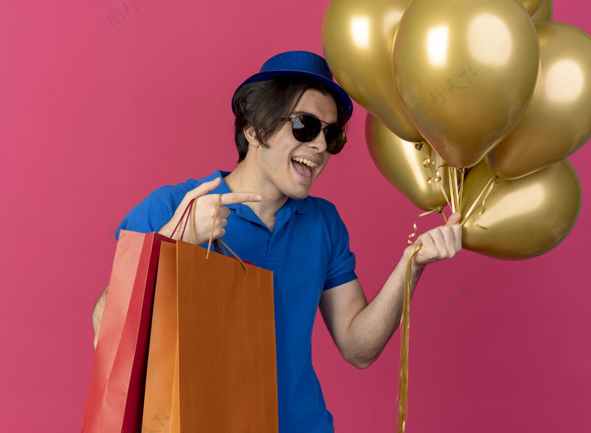 复制一个戴着太阳眼镜 戴着蓝色派对帽 手里拿着氦气球和纸购物袋的快乐帅哥氦太阳男人
