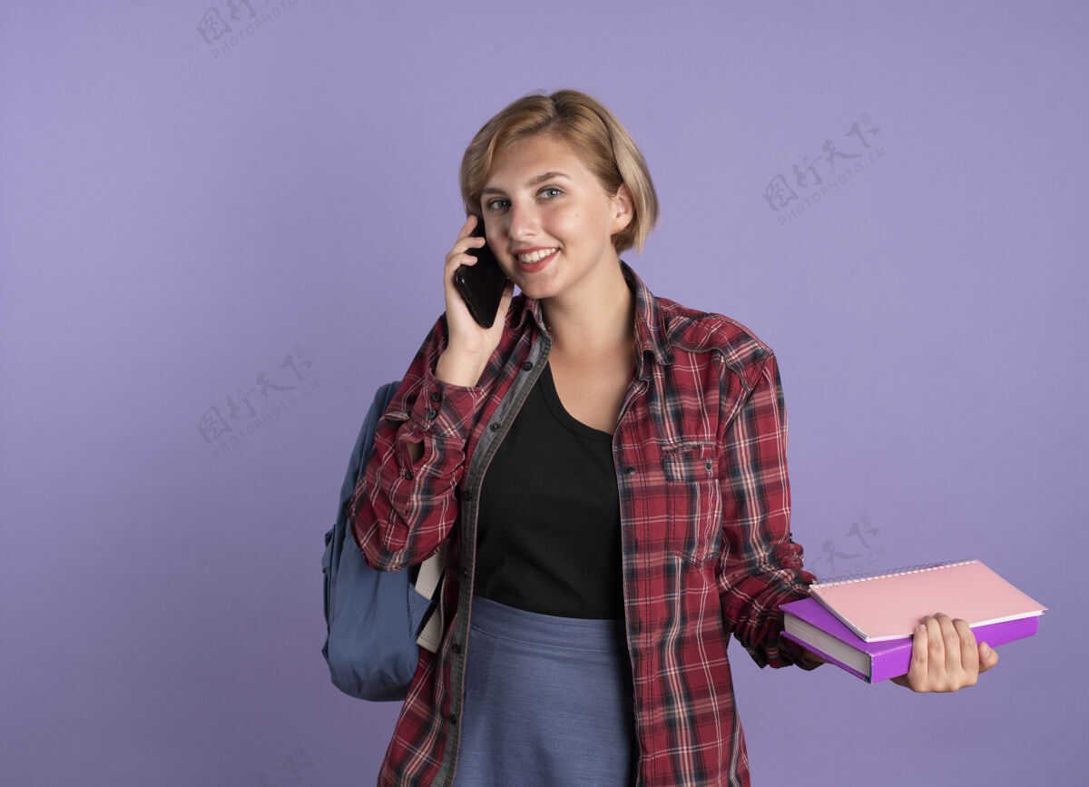 微笑微笑着的年轻斯拉夫学生女孩背着背包拿着书和笔记本在电话里交谈笔记本斯拉夫紫色