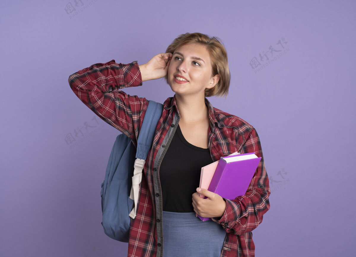 紫色高兴的年轻斯拉夫学生女孩戴着背包 手放在头上 拿着书和笔记本看着身边年轻背包空间