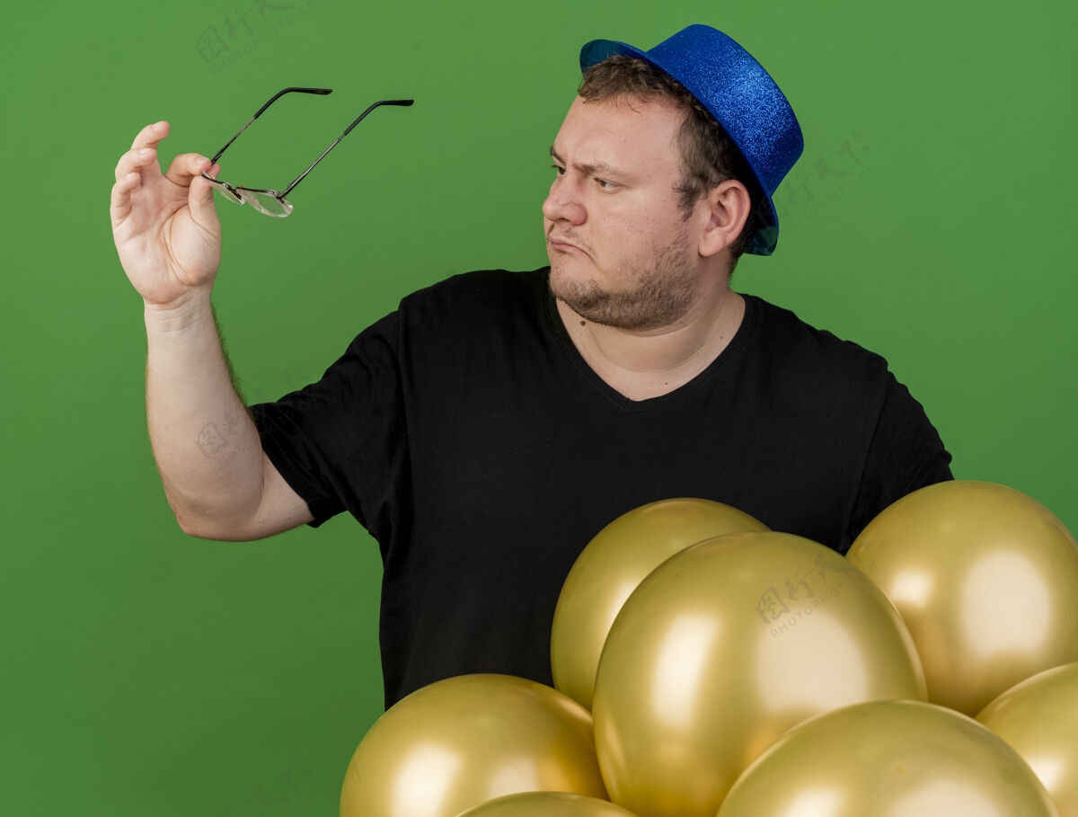 蓝色不知所措的成年斯拉夫男子拿着眼镜看着戴着蓝色派对帽站着的氦气球举行气球成人