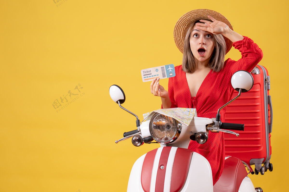 额头迷惑不解的穿着红衣服的年轻女士拿着车票 手放在她的额头上 骑着轻便摩托车红人表情