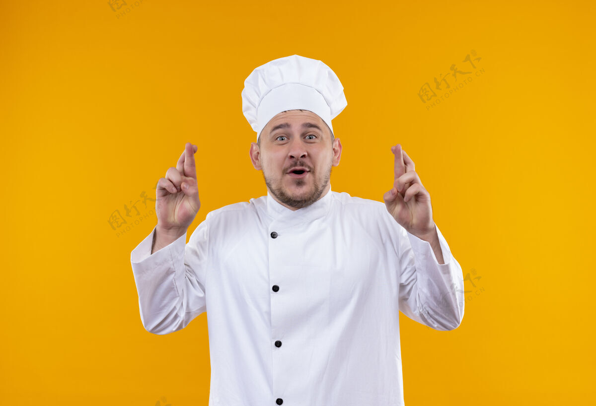 年轻印象深刻的年轻帅哥厨师在厨师制服做交叉手指手势隔离在橙色的墙上手指手势厨师
