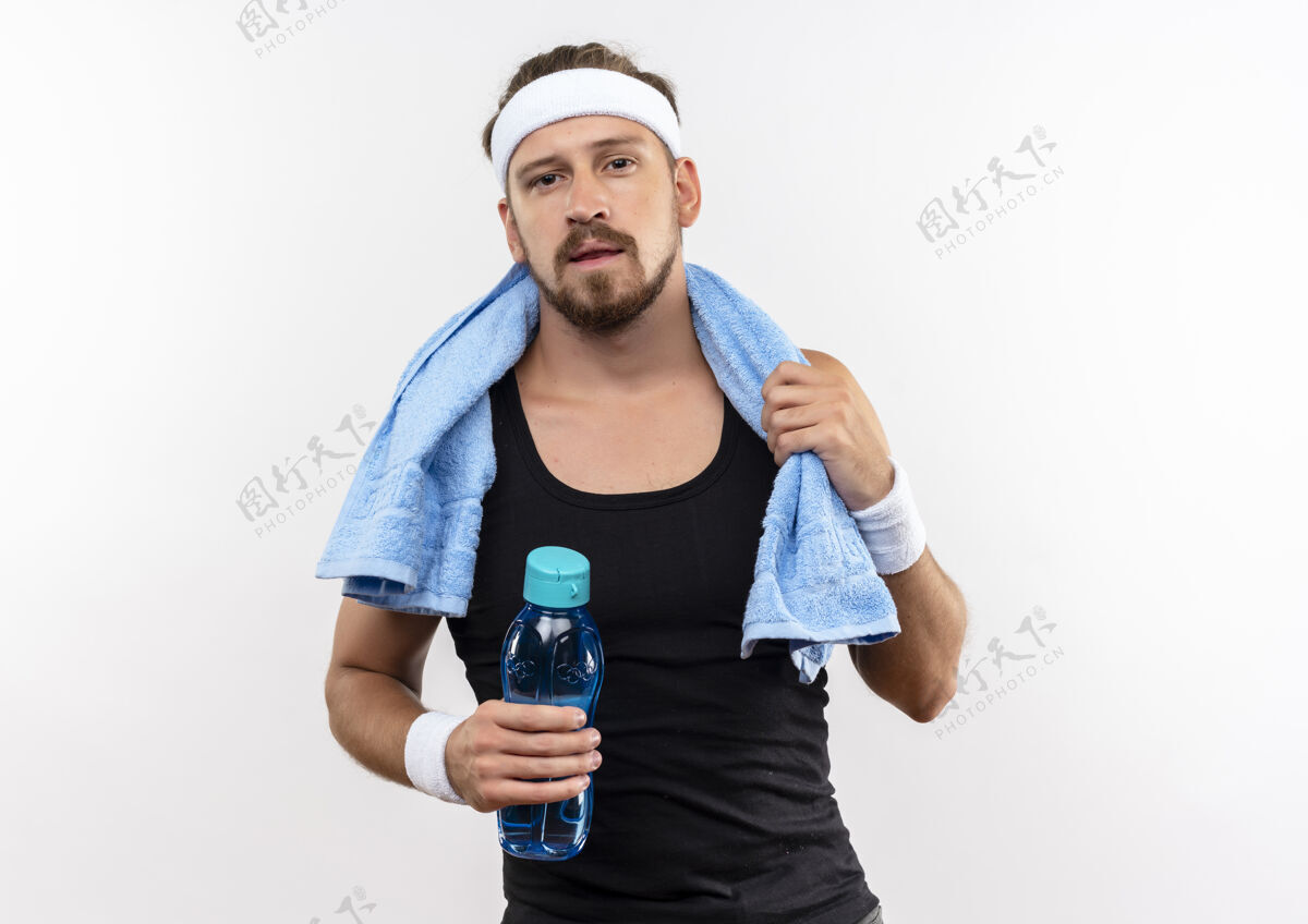 自信自信的年轻帅气的运动型男人戴着头带和腕带 脖子上围着毛巾拿着水瓶和毛巾隔离在白墙上腕带头带脖子