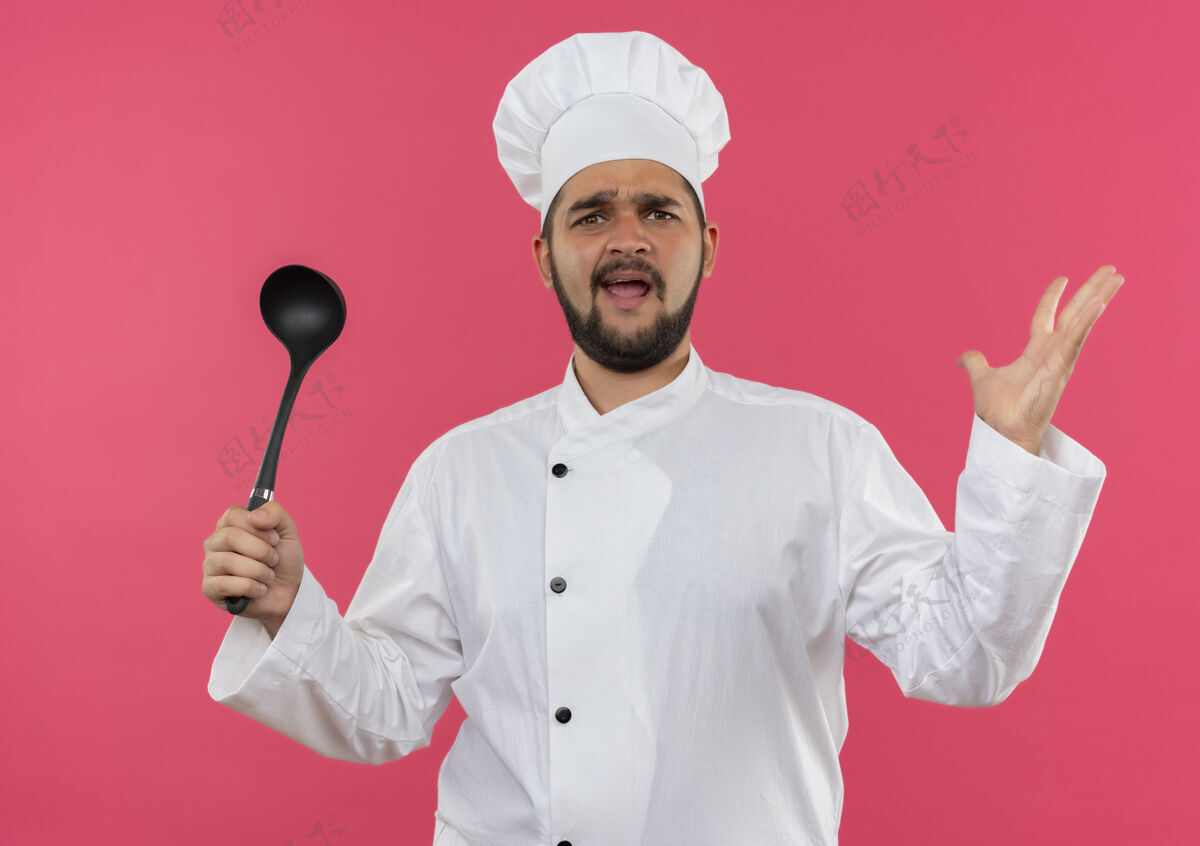制服恼怒的年轻男厨师穿着厨师制服 手里拿着勺子 在粉红色的墙上孤立着一只空手手年轻勺子
