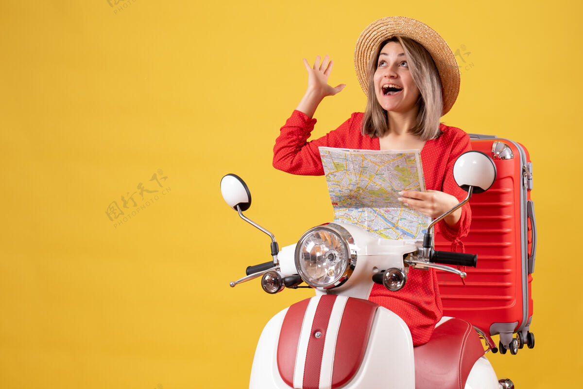 坐着骑着轻便摩托车的漂亮女孩拿着红色的手提箱 举着地图向上看时尚红色地图