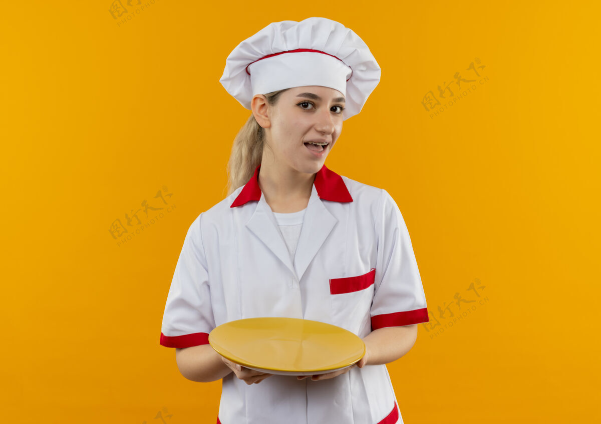 空年轻漂亮的厨师穿着厨师制服 戴着牙套 把空盘子隔离在橙色的墙上牙科漂亮拿着