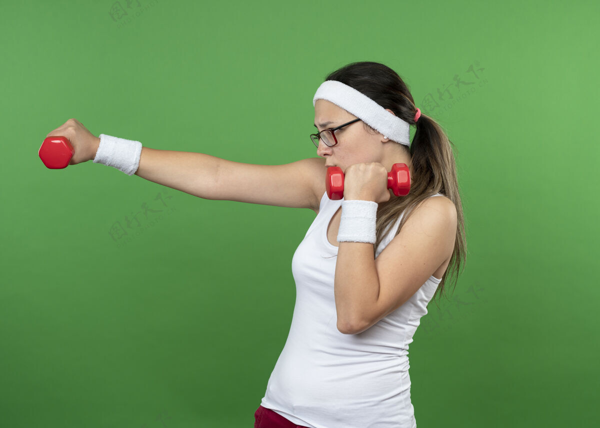 光学自信的年轻运动女孩戴着眼镜戴着头带和腕带举着哑铃年轻运动头带