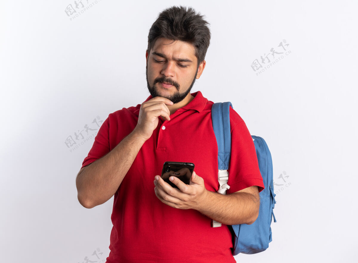 年轻年轻的留着胡子的学生 穿着红色马球衫 背着背包 拿着智能手机 站在那里困惑地看着它胡子抱背包