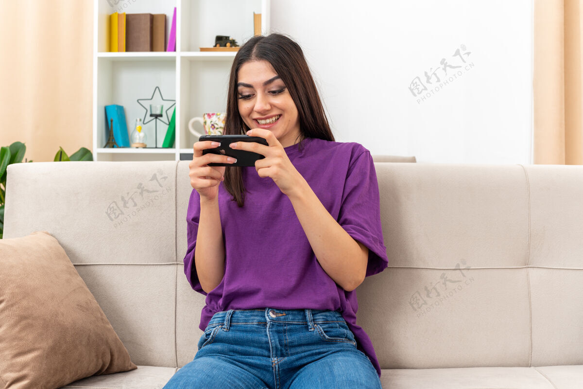 坐穿着休闲服的小女孩用智能手机玩游戏快乐而兴奋地坐在明亮的客厅沙发上休闲玩衣服
